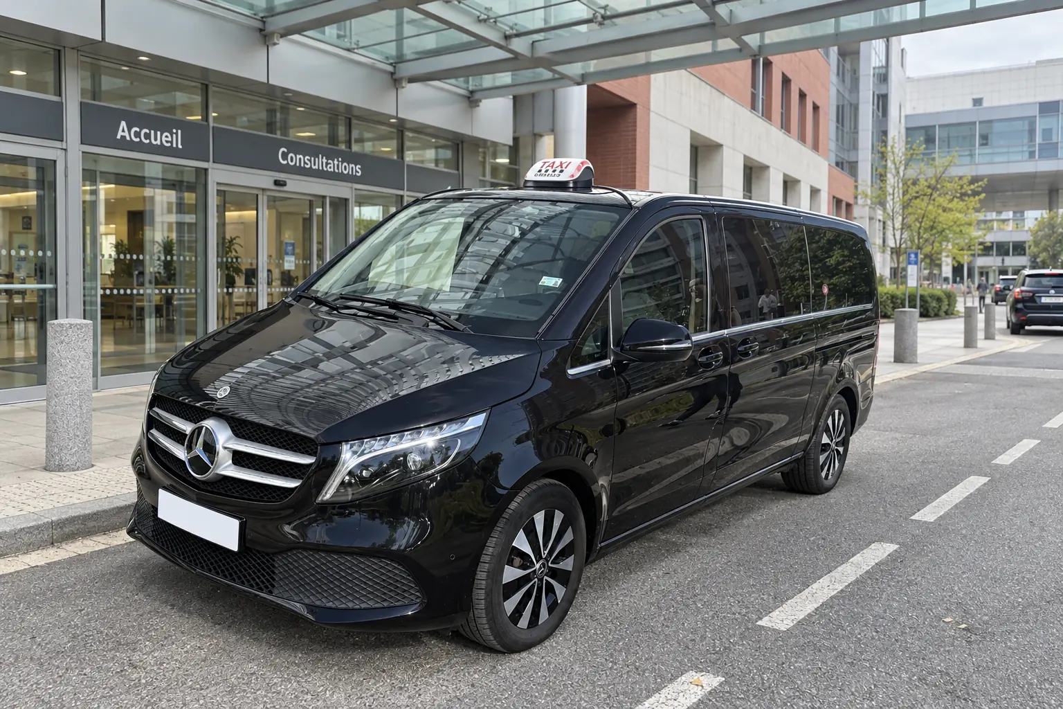 La Classe V noire de VK Taxi arrêtée devant l’entrée vitrée d’un grand hôpital, dans la voie de dépose