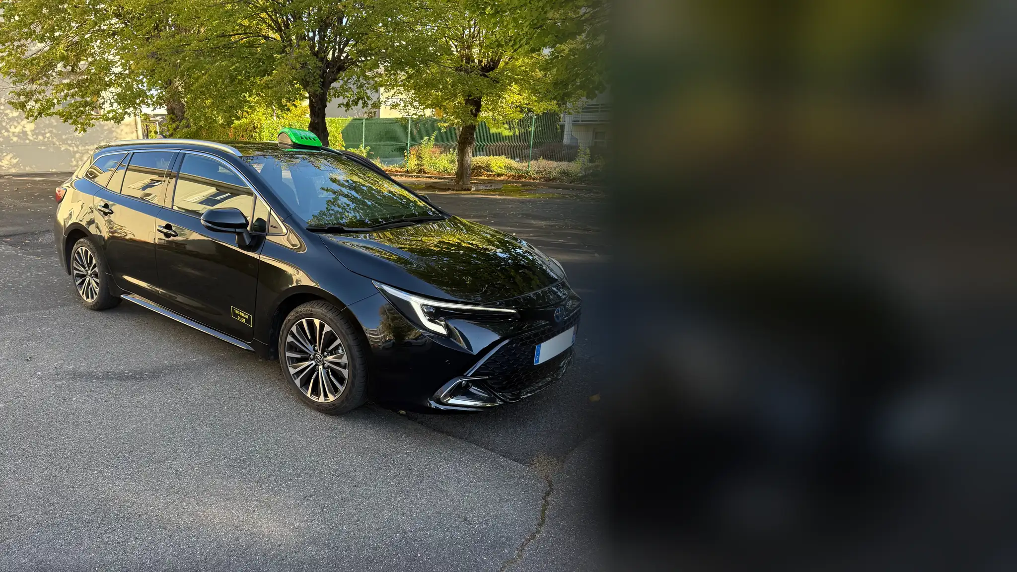 Le taxi relais de VK Taxi et Services, une Toyota Corolla break hybride noire, lumineux TAXI vert sur le toit, garée sous les arbres