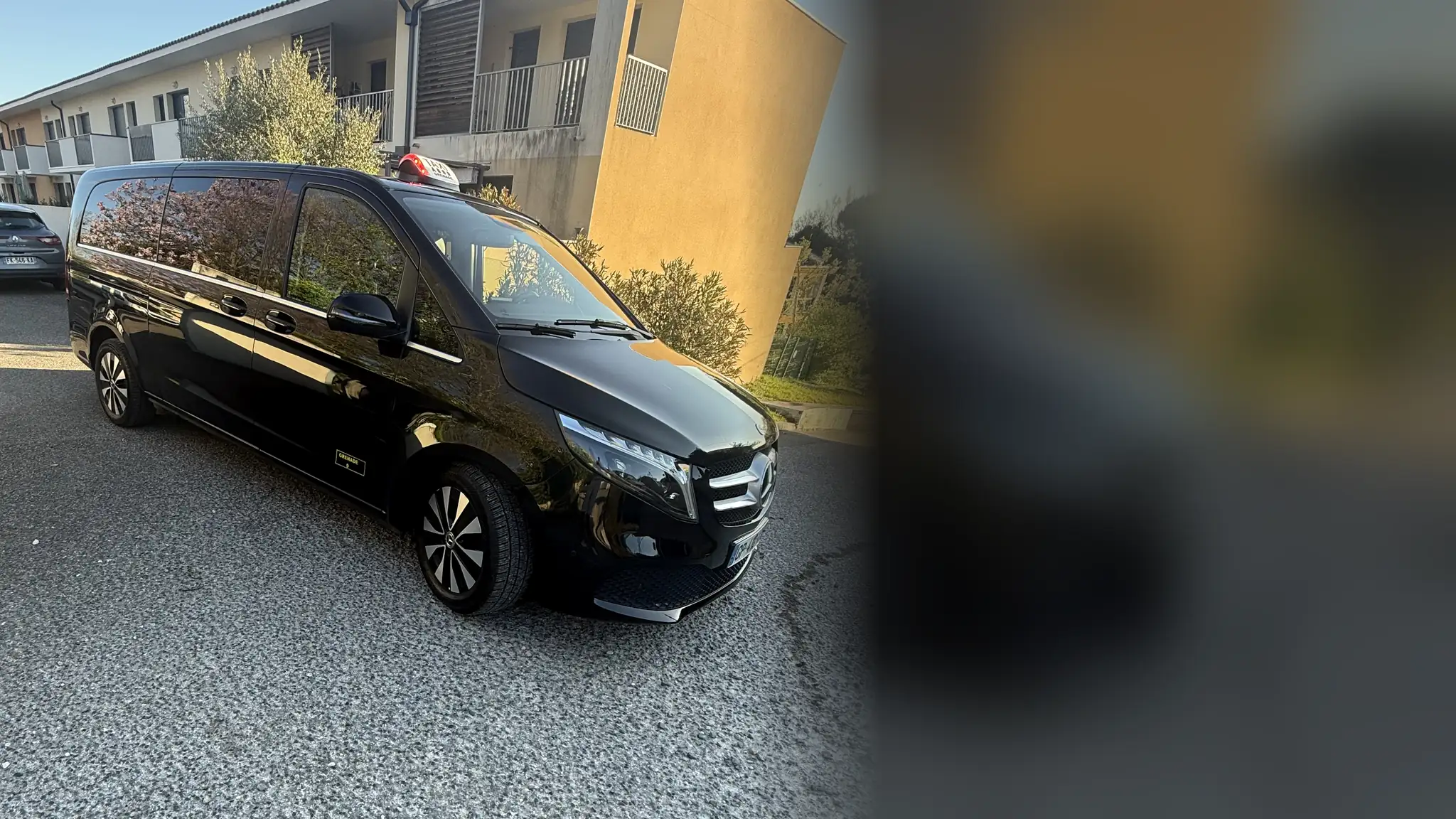 Le Mercedes Classe V noir de VK Taxi et Services, lumineux TAXI sur le toit, garé devant une résidence au soleil